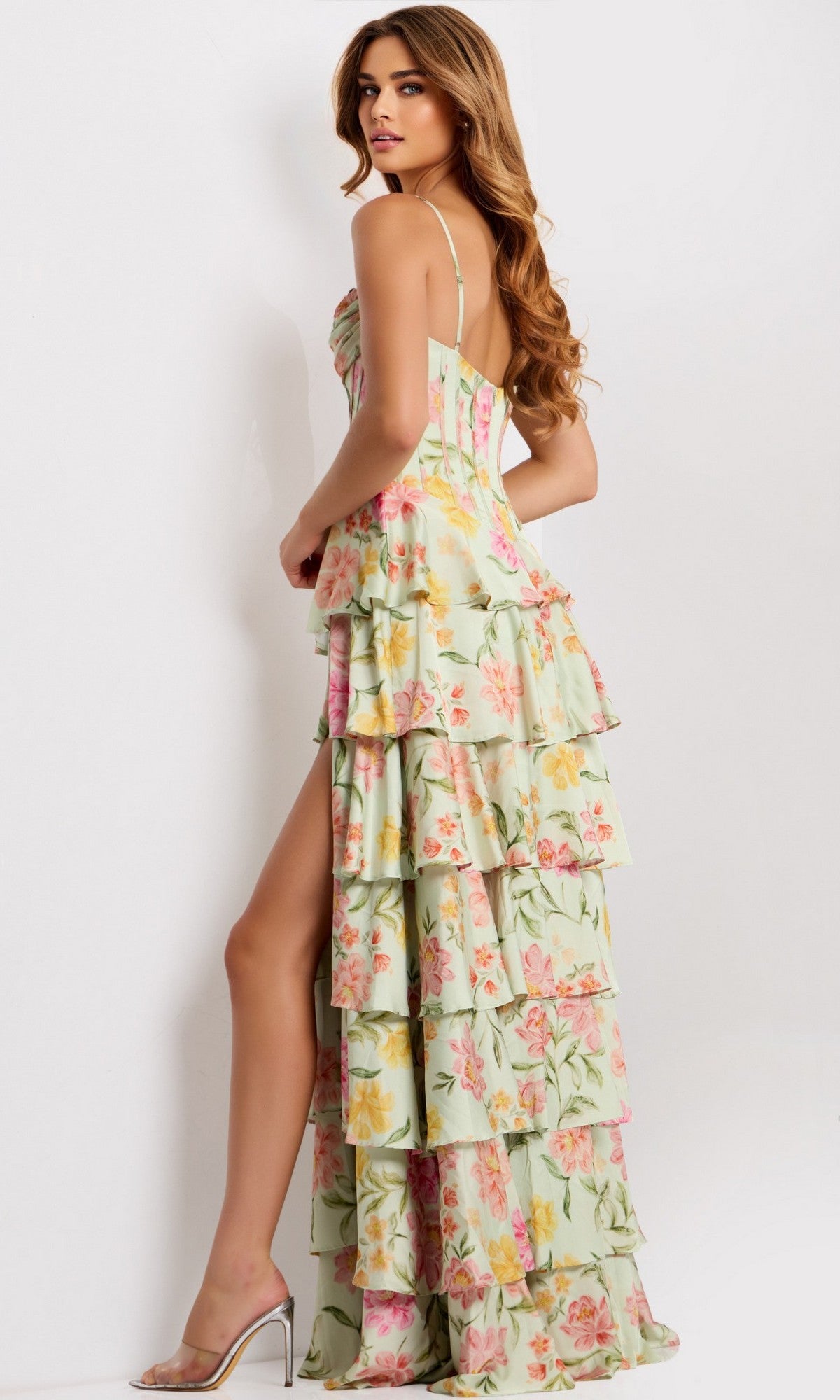 A woman models the Long Prom Dress: Jovani 47561, which features a floral print, corset bodice, ruffle skirt, and thigh-high slit. She pairs it with strappy high heels and poses against a plain white background, looking over her shoulder.